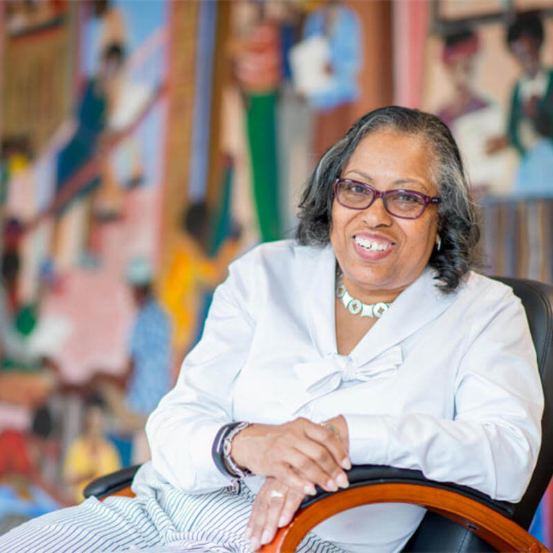 Dr. Gourdine sits in a chair, facing the camera with a colorful mural behind her